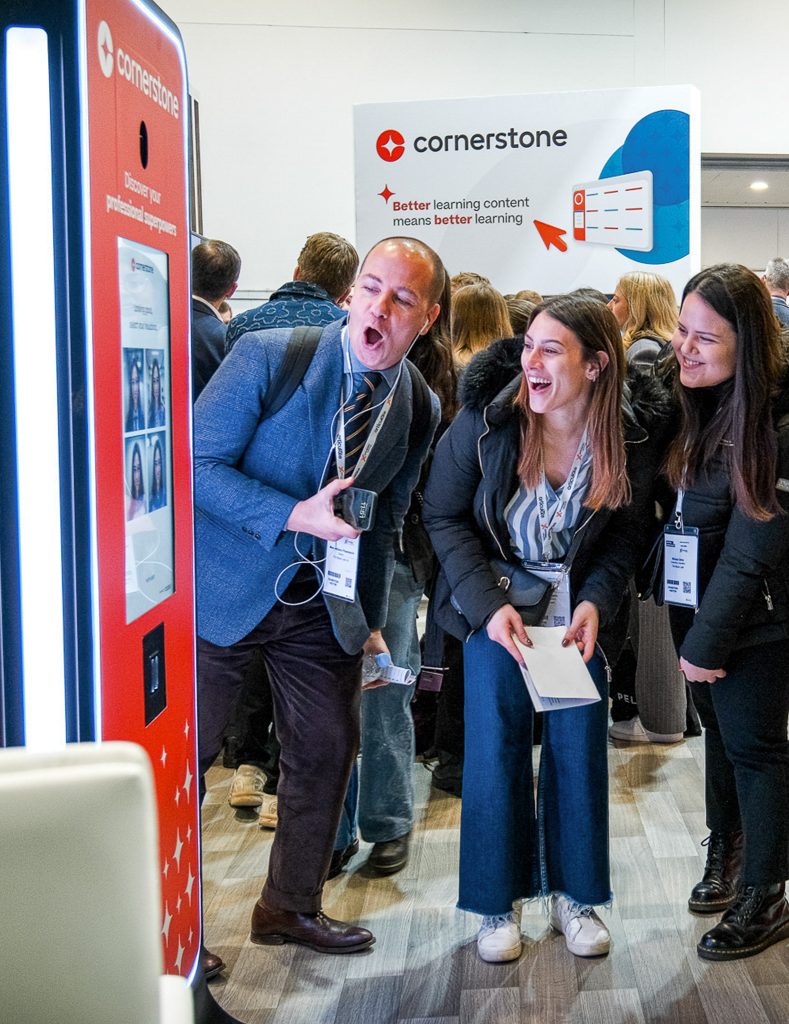 Guests reacting with excitement to AI Photo Booth outputs at the Cornerstone stand during the Learning Technologies trade show.